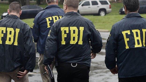 Four men walk away from the camera wearing dark jackets with bright yellow 'FBI' letters on the back, in a parking lot with vehicles visible.