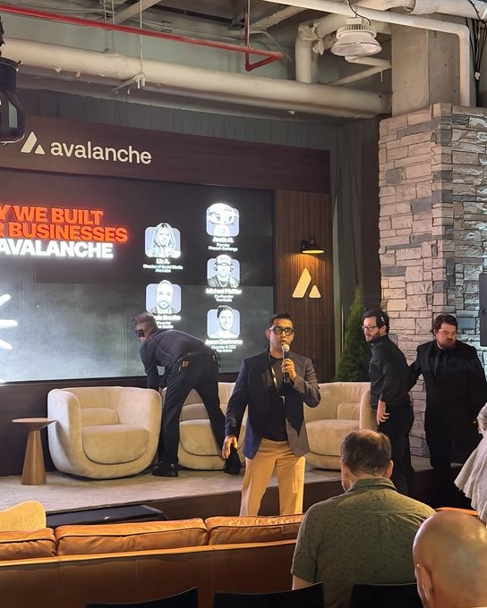 A man in a suit speaking on stage at an Avalanche event with a screen showing 'WHY WE BUILT THE BUSINESSES AT AVALANCHE' and team member profiles.