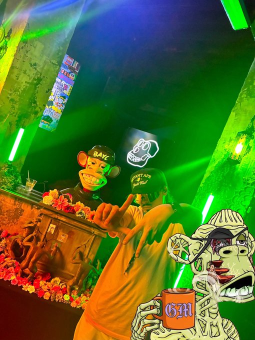 A photo of two people at what appears to be a bar or club, wearing Bored Ape and Rantape NFT hats, overlaid with an illustration of a zombie-like ape character holding a mug labeled 'GM' and smoking.