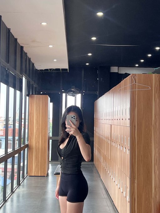 A woman takes a mirror selfie in what appears to be a locker room or changing area, wearing a black fitted top and shorts while holding up her phone to capture the image. The setting, with rows of wooden lockers and large windows, contrasts with her caption positioning herself as a 46-year-old $HYPE accumulator from Singapore engaging with the Hyperliquid ecosystem, likely at a fitness or wellness facility.