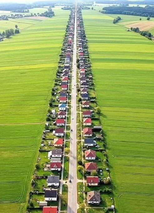 Aerial photograph of a remarkably straight, narrow village street lined with houses on both sides, cutting through vast green agricultural fields stretching to the horizon.