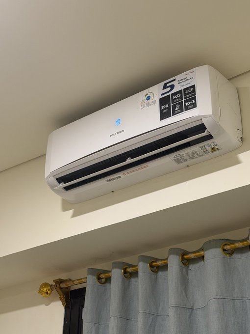 A white Polytron brand split-type air conditioner mounted on a wall above gray curtains, displaying a '5 Year Warranty' sticker and energy rating labels including '380', 'R32', and '10+3'.