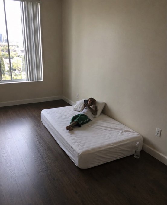 A person lies on a bare mattress in an otherwise empty room with hardwood floors and a large window, looking at their phone.