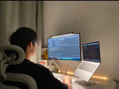 A person sitting at a desk working on a laptop alongside an external monitor displaying code.