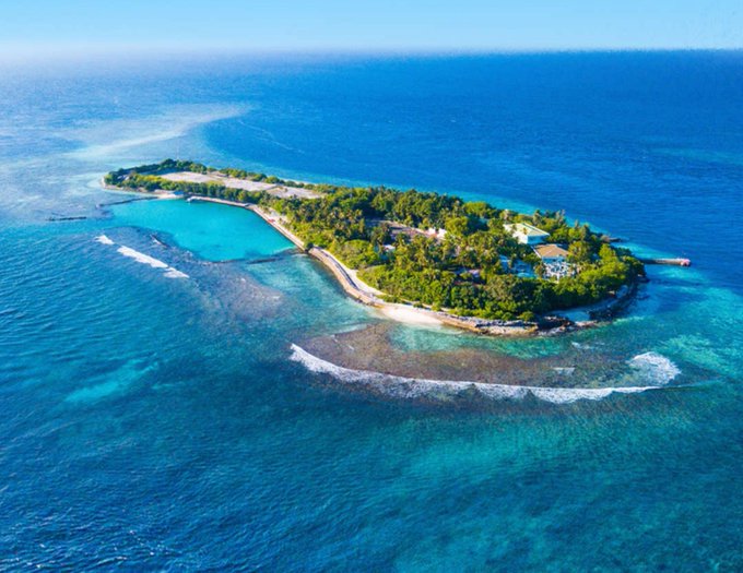 An aerial photograph of a small, lush green tropical island surrounded by clear blue and turquoise ocean waters, with a few buildings visible among the trees.
