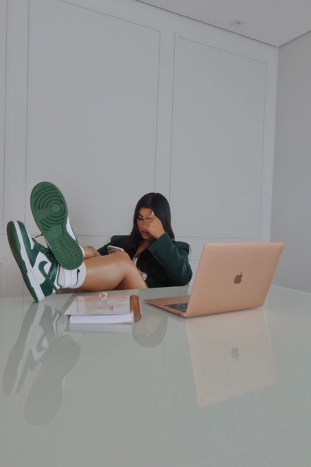 A woman with long dark hair wearing a green blazer and green/white sneakers reclines with her feet up on a glossy white table, looking at her phone next to an open rose-gold Apple MacBook.