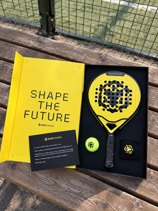 BNB Chain 'Shape the Future' promotional package on a wooden bench, containing a yellow booklet, a custom padel racket labeled 'Build N Build', a tennis ball and an info card.