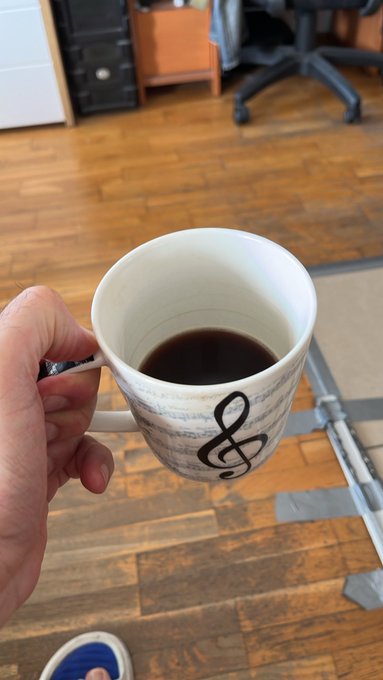 A hand holding a white ceramic mug filled with black coffee, decorated with a treble clef musical note and sheet music design, with a wooden floor and office chair visible in the background.