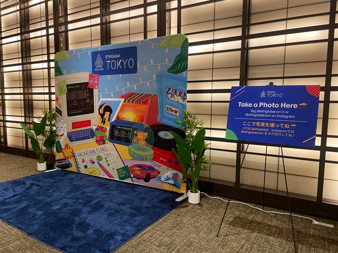 An indoor exhibition space featuring a large decorative photo standee with arcade and travel themes, alongside a blue sign on an easel reading 'ETHGlobal TOKYO - Take a Photo Here' with an Instagram handle and Japanese text.