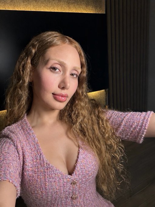 A young woman with long, curly blonde hair wearing a pink and lavender tweed top poses for a selfie in a dimly lit setting. The caption "GM crypto enjoyers" suggests she's greeting the crypto community with a cheerful morning message.