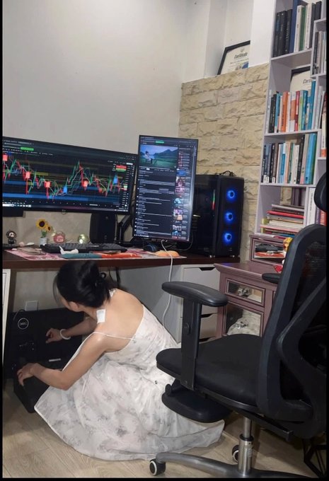 A young woman in a white dress kneeling on the floor working on a desktop PC tower beneath a dual-monitor setup displaying trading charts, with a bookshelf and sunflower decor in the background.