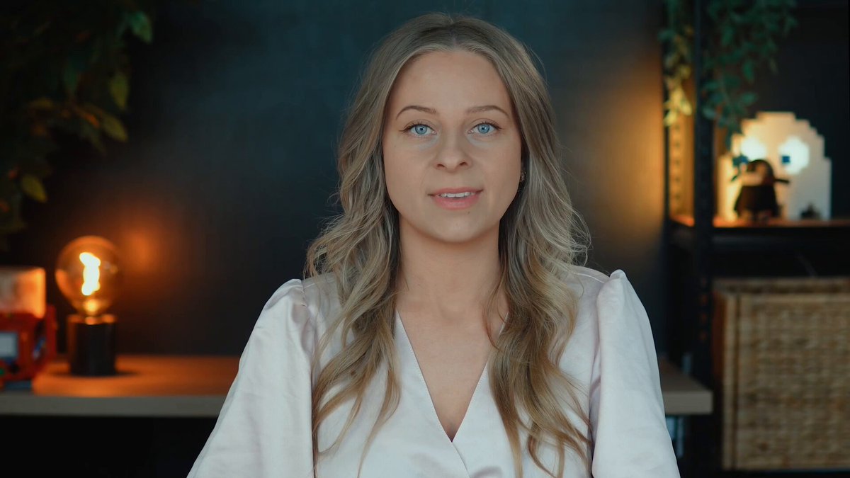 The image shows a woman with blonde hair wearing a white blouse sitting at a desk or table. She has her eyes closed, giving an impression of deep thought or contemplation. The background features a dark blue wall that contrasts with the light brown wooden desk in front of her. There is also a lamp on the desk, which adds to the overall ambiance of the scene.