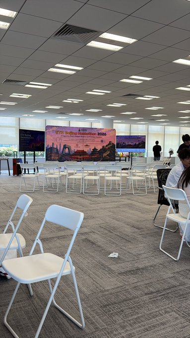 The image shows a conference room with white chairs arranged around a large screen displaying an advertisement for Bitcoin. The room has a gray carpet, and there are several people sitting at tables or standing near the screen. Some of them appear to be engaged in discussions or presentations related to Bitcoin.