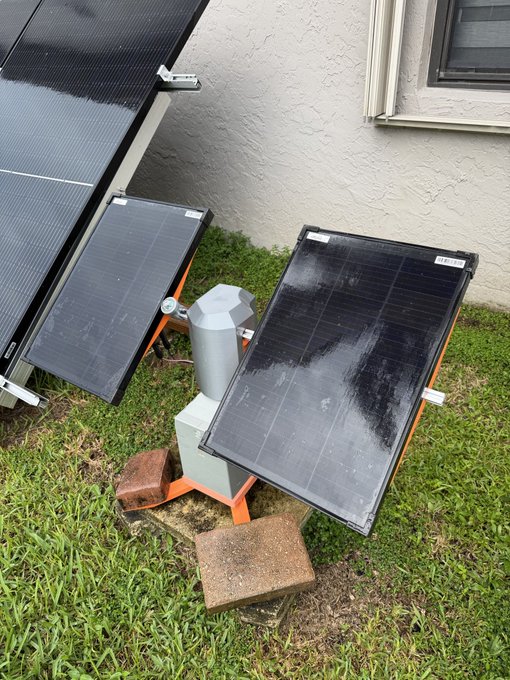 Two solar panels and a metallic device are set up on the grass next to a house wall, propped up on bricks and an orange stand.