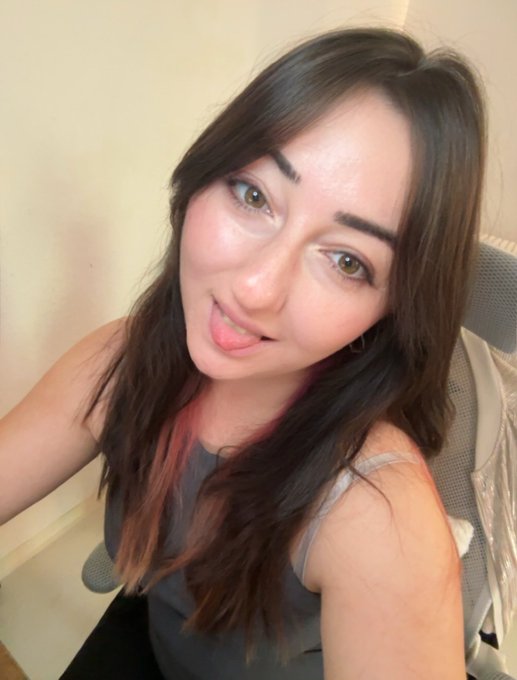 A selfie-style photo of a young woman with long dark hair with reddish ends, wearing a gray tank top, looking at the camera against a plain background.
