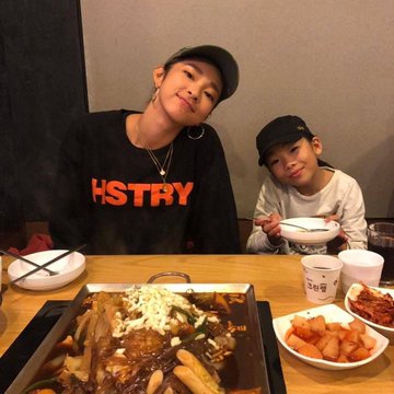 Two people sit at a wooden table in a restaurant, with the person on the left wearing a black sweatshirt with "HSTRY" in red text, while a hot dish, fried chicken, and side dishes are spread on the table.