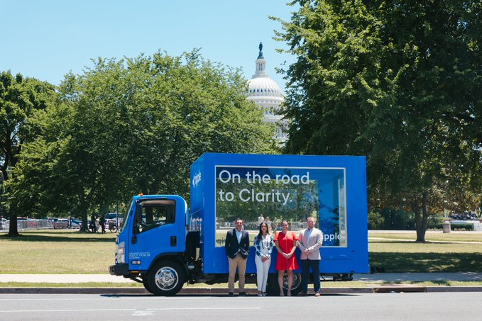 A bright blue branded truck displaying "On the road to Clarity" and the Ripple logo is parked on Capitol Hill, where four people stand in front of it with the U.S. Capitol dome visible in the background, illustrating Ripple's lobbying campaign urging lawmakers to pass the Clarity Act.