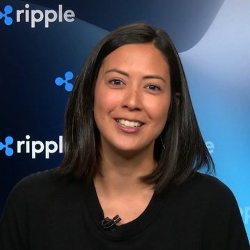 The image features a woman with short hair wearing a black shirt, smiling at the camera. The background of the image includes a blue banner that says "Ripple" and "RLUSD", suggesting that she might be discussing Ripple cryptocurrency or related events. The caption in the image reads "Monica Long says institutional demand for $XRP and RLUSD is unlike anything we've seen." This implies that Monica Long has made some statement about Ripple cryptocurrency, possibly expressing her opinion on its value or importance.