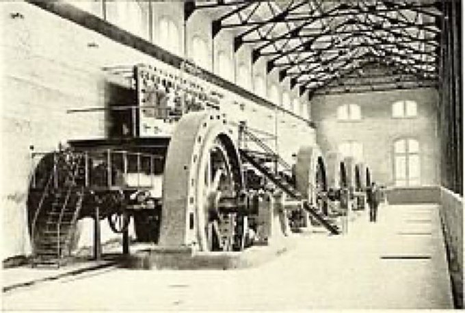 A vintage black and white photograph shows large industrial machinery with wheels and panels inside a high-ceilinged building with arched windows.