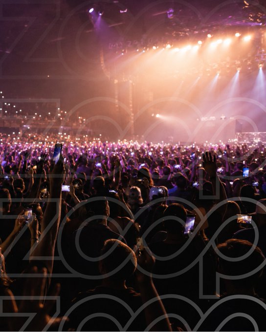 A crowded concert arena lit with pink and orange stage lights, with fans holding up phones to record the performance.