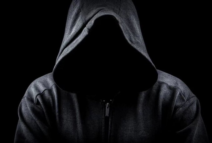 A dark, shadowy figure wearing a hooded sweatshirt stands against a black background, with their face completely obscured.