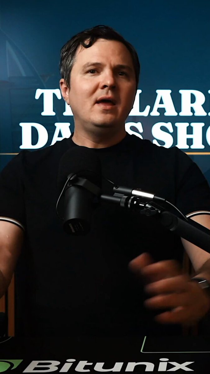 The image features a man wearing a black shirt sitting behind a microphone at a table with a computer monitor. The man appears to be giving a speech or presentation, possibly related to Bitcoin or cryptocurrency. The background of the image includes a blue wall with white text that reads "The Digital Labels Show".