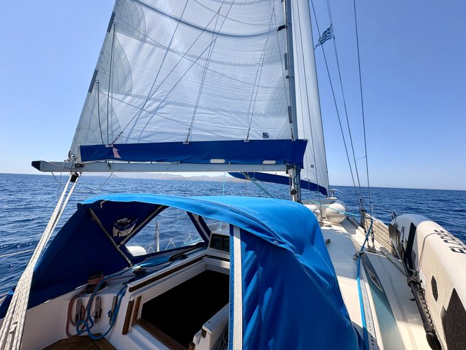 A sailboat glides across calm blue waters, its white mainsail and blue canvas bimini top catching the sunlight. A small Greek flag flutters from the rigging, complementing the post's "GM Legends!" celebration of sailing heritage.