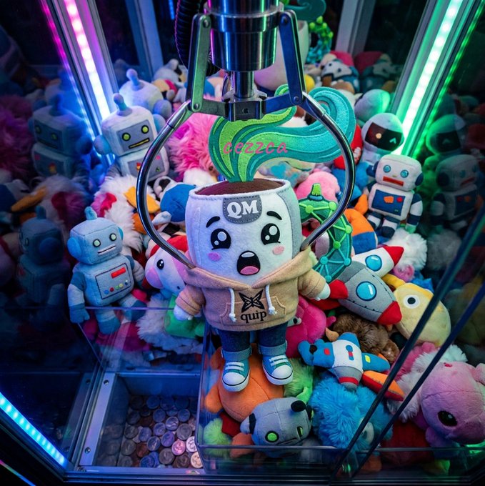 A claw machine filled with various plush toys including robots and colorful characters, with the mechanical claw grasping a mug-shaped plush toy labeled 'QM' that has green leaves on top.