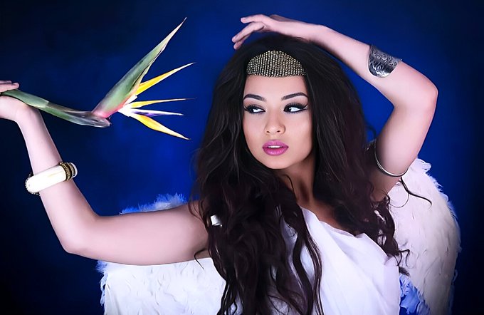 A woman with long dark hair wearing a white feathery outfit and decorative headpiece poses against a deep blue background while holding a bird of paradise flower near her face.
