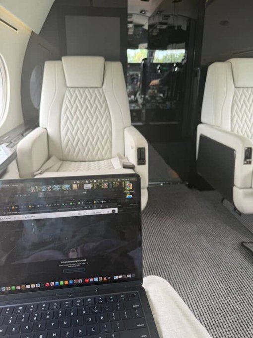 A view inside a private jet showing cream-colored leather seats with a person's laptop displaying a dark-themed web page in the foreground.
