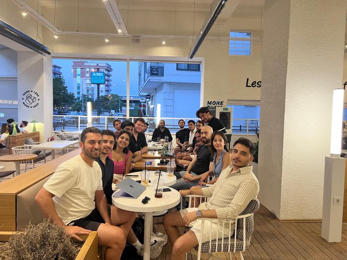 A group of around a dozen attendees poses together at a casual indoor venue during the Stellar Türkiye Meetup in İzmir, with a laptop on the table and "KOMÜN" and "Les More" signage visible in the background. The relaxed gathering reflects the post's theme of connecting with builders, developers, and the local Web3 community within the Stellar ecosystem.