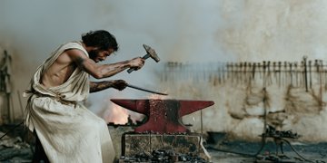 A man in a sleeveless white garment forges metal on an anvil with a hammer in a smoke-filled workshop, reminiscent of a blacksmith scene.