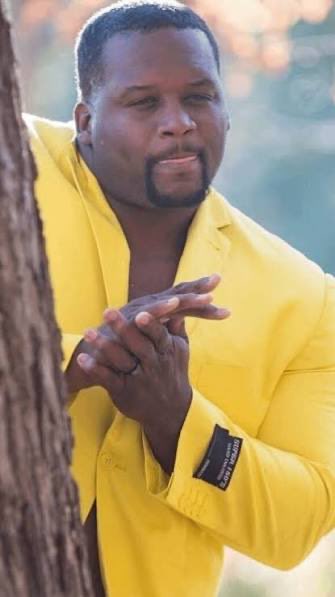 A man wearing a bright yellow blazer poses with a hand-to-chin gesture while leaning against a tree. The image captures a confident, contemplative moment that matches the post's mention of @wagmiAlexander from @AerodromeFi, styled like a candid celebrity "live look."