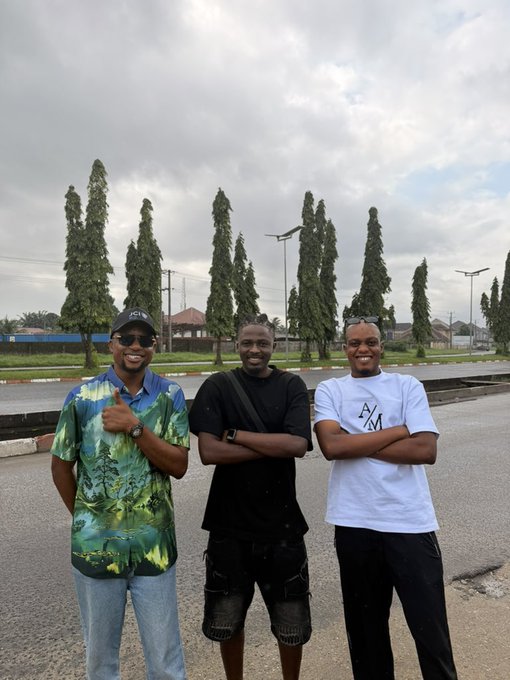 A photograph of three smiling men standing together outdoors on a paved road, with tall cypress trees and an overcast sky in the background.