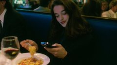 A still from a film showing a young woman in a dimly lit restaurant, looking down at her phone while holding food.