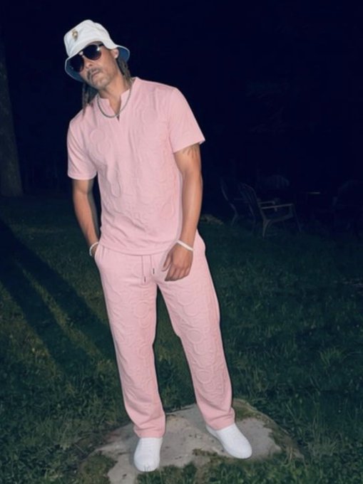 A man wearing a white bucket hat, sunglasses, and a pink textured t-shirt and matching sweatpants set stands on grass at night.