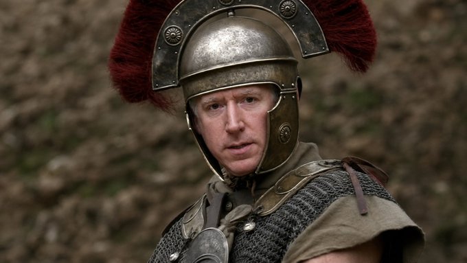A man wearing a Roman centurion helmet with a red horsehair crest and chainmail armor looks off to the side with a serious expression.