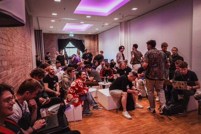 A crowded indoor event space with exposed brick walls and pink/purple ceiling lighting, where many young people are gathered with laptops, sitting on white blocks or standing.