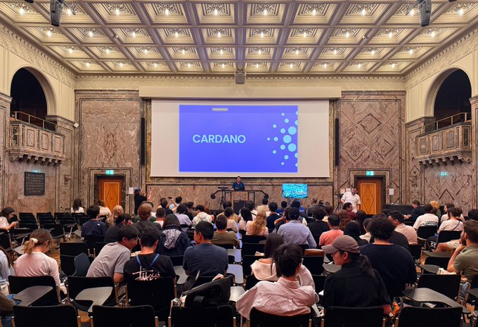 The image shows a presentation on Cardano taking place at the University of Zurich (UZH), with a speaker on stage addressing a large audience of students seated in an ornate lecture hall, illustrating the blockchain event described in the accompanying post.