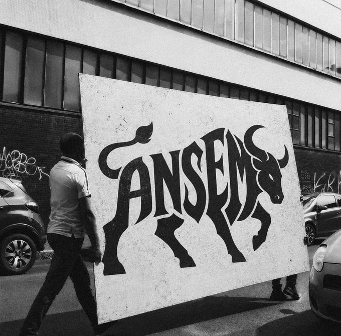 A person carries a large sign featuring a black bull graphic and the bold text "ANSEM" down a city street, visually representing the post's boast that $ANSEM has already built the attention, distribution, and community most memecoins spend their existence chasing.