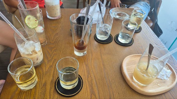 A wooden table at a café set with multiple half-empty glasses of iced coffee, water, and other drinks with straws, suggesting a group hangout that has recently wrapped up.