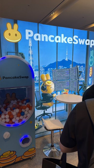 A high-rise office features a 'PancakeSwap' themed area with a rabbit mascot figurine, a gachapon-style machine filled with balls, and a large branded sign, overlooking a city skyline at dusk.
