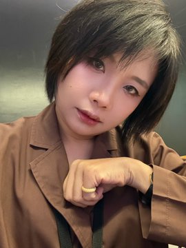 A woman with short brown hair and bangs wearing a brown blazer poses for a selfie, with her chin resting on her hand.