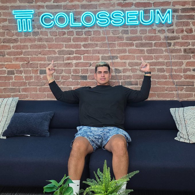 A man sits on a dark blue couch against a brick wall featuring a bright neon "COLOSSEUM" sign, pointing up with both index fingers in an enthusiastic pose. The post's "GM San Fran" greeting and mention of the Colosseum office's "unreal energy" align with the image, showcasing the Colosseum brand's vibrant headquarters and conveying the exciting, shipping-focused atmosphere of their Solana-focused work.