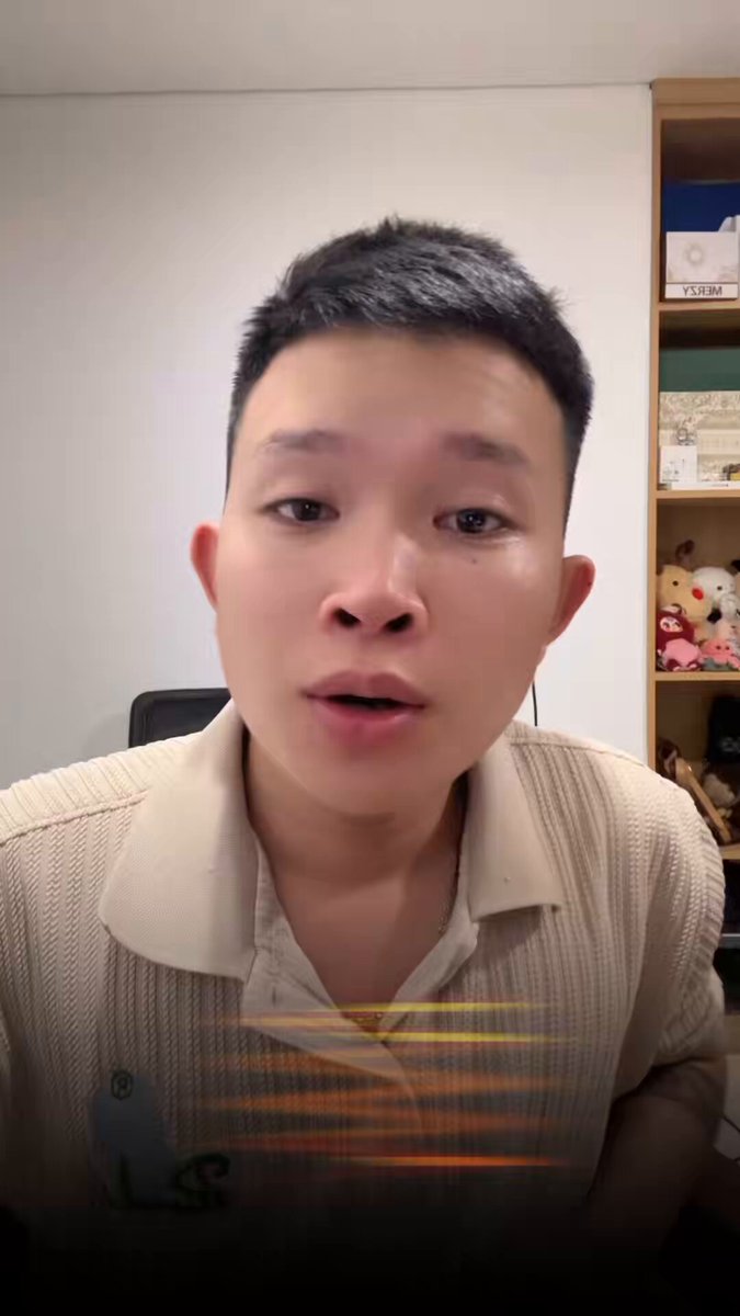 The image shows a man wearing a beige sweater, with his face partially obscured by the camera lens. He appears to be making a funny or serious facial expression as he looks at the camera. The background of the photo features a white wall, which contrasts with the man's attire and adds depth to the scene. There are also several stuffed animals scattered around the room, adding an element of whimsy to the image.