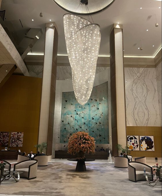 The accompanying post "gm lets cook" features a crypto-trading slang expression often used by traders to signal readiness for market action, which connects to the image's depiction of a luxurious hotel lobby with a grand crystal chandelier, elegant seating, and modern artwork—evoking the aspirational, high-stakes lifestyle and excitement associated with successful crypto trading.