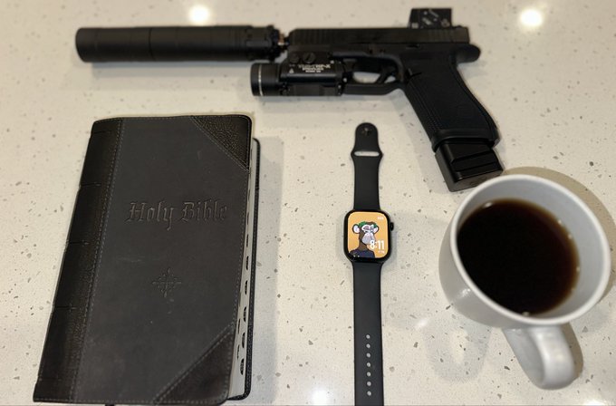 A flat lay arrangement on a white speckled counter showing a handgun with a suppressor and laser, a black 'Holy Bible', an Apple Watch with a cartoon dog face, and a white mug of black coffee.