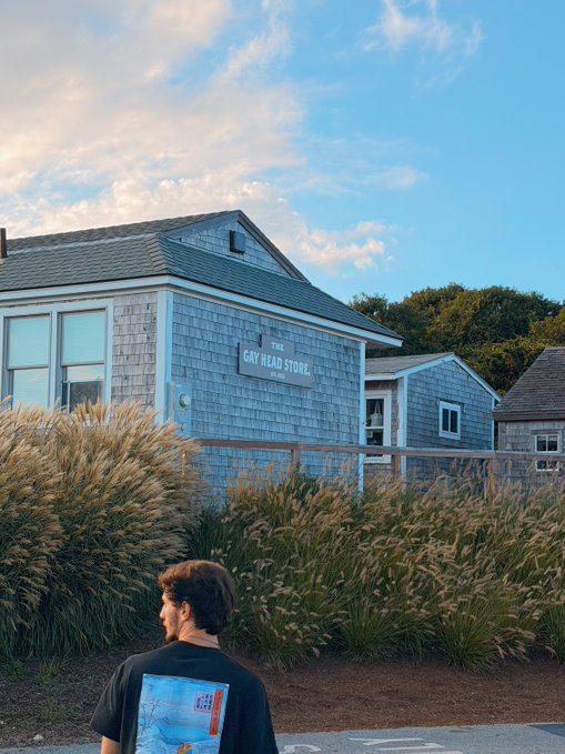 A young man walks past a gray shingled building labeled "THE GAY HEAD STORE" with tall grass swaying in the foreground under a partly cloudy sky, seemingly a moment of calm reflection during a profitable Ethereum ($ETH) trading day.
