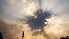A dramatic sky photograph showing sun rays piercing through clouds, with silhouettes of utility poles and tree tops visible in the foreground.