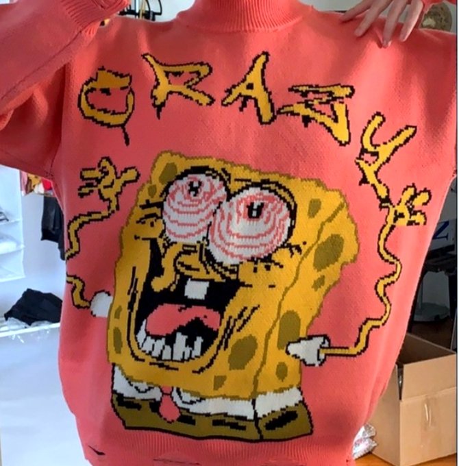 A coral-red knitted sweater is held up, featuring a large, pixelated cartoon design of a wide-eyed SpongeBob SquarePants with the word 'GRAVITY' stylized above him.